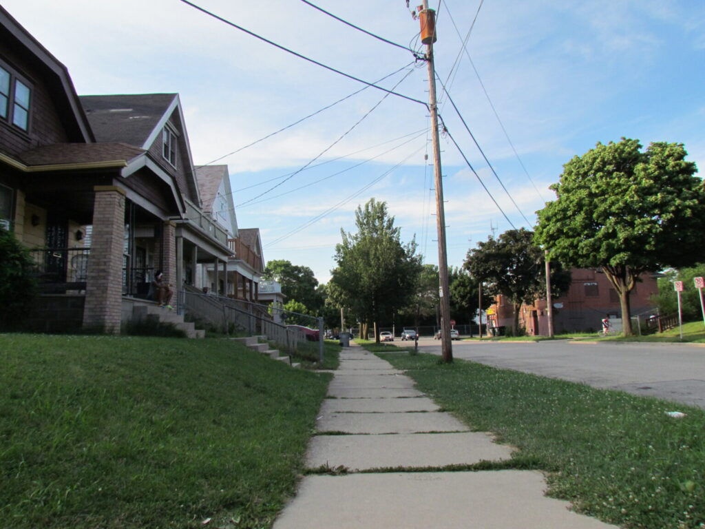 An image of a sidewalk, green grass, power lines crossing overhead, and houses on the left.